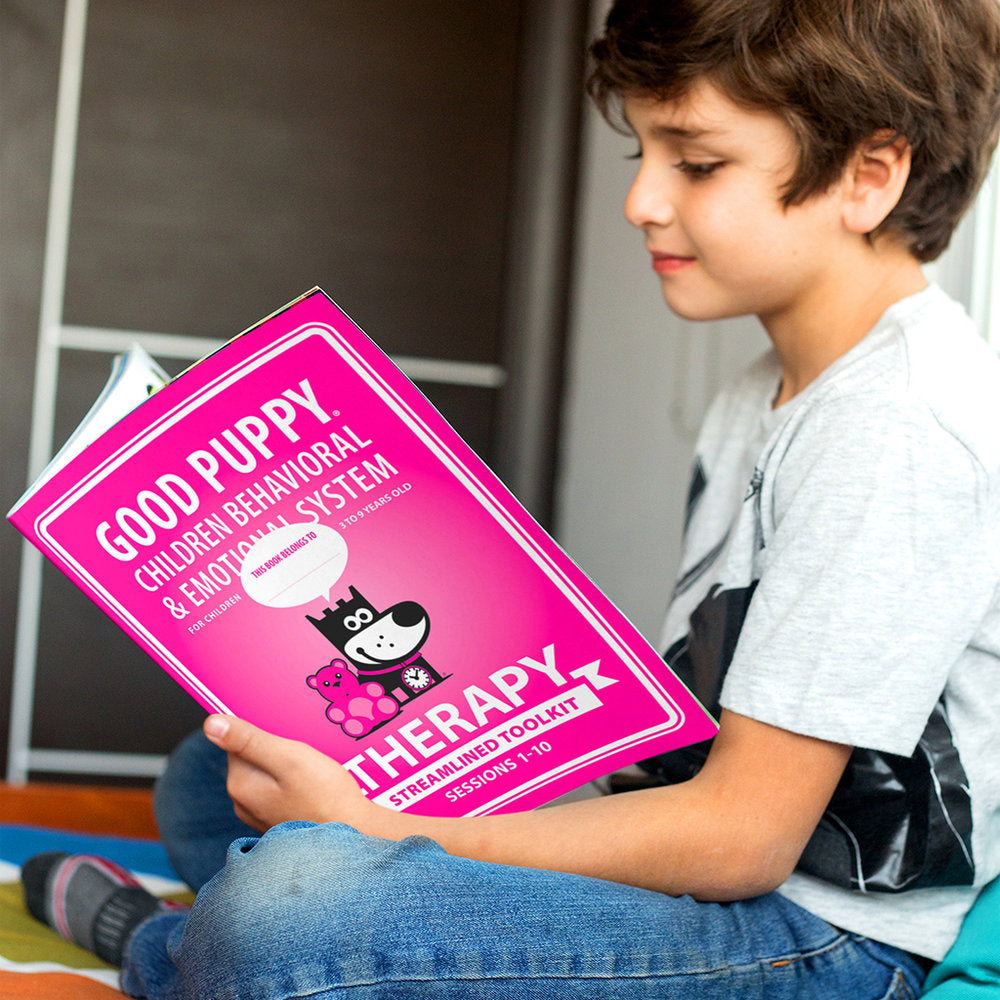 A young boy is sitting on the floor, reading a book titled "Good Puppy Children Behavioral & Emotional System: Therapy Streamlined Toolkit, Sessions 1-10." The book has a pink cover featuring a cartoon dog and teddy bear. The boy appears focused and engaged with the content.