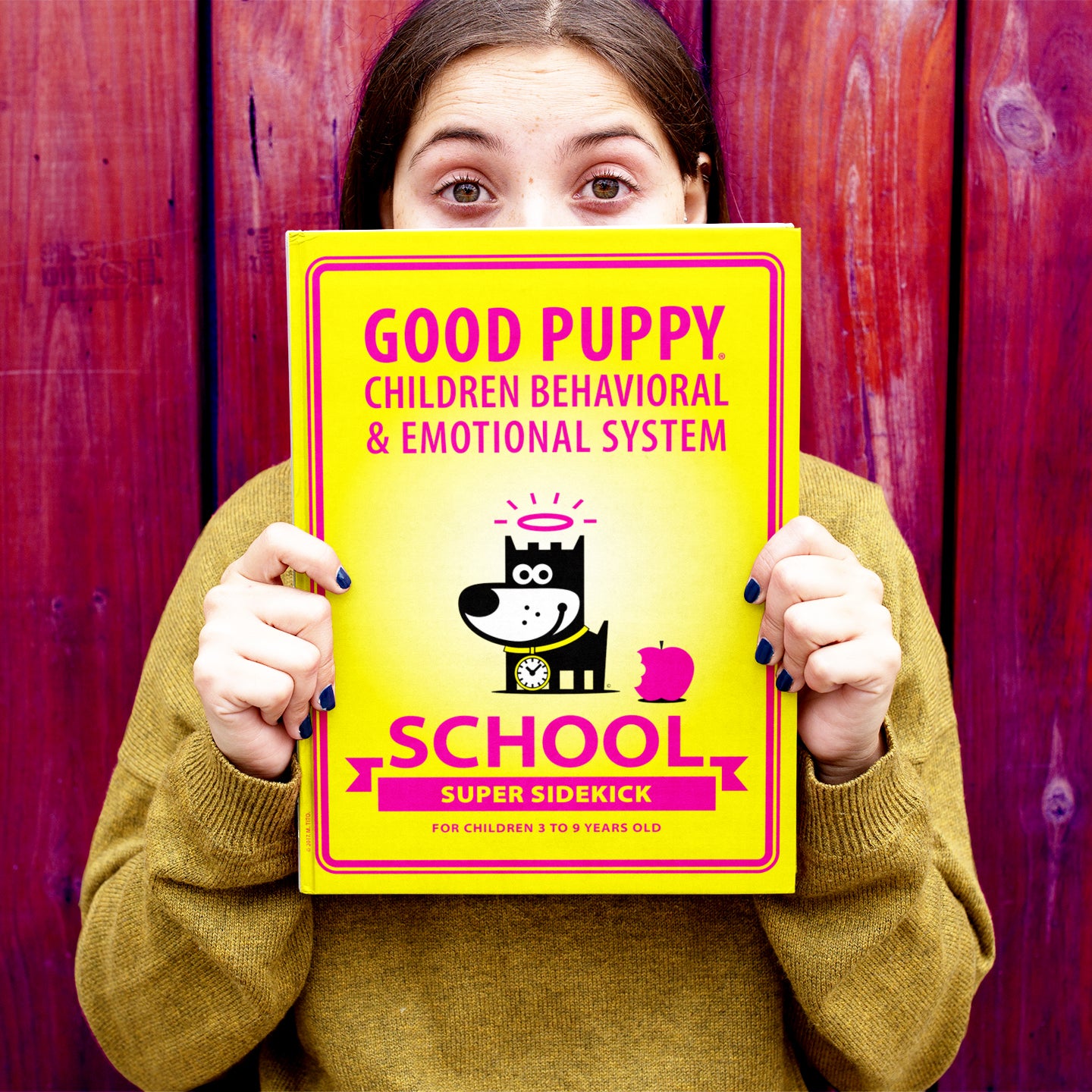 The image shows a person holding a yellow book for the "Good Puppy Children Behavioral & Emotional System" for school. The book features a cartoon dog with a halo and a clock collar, along with a bitten apple. The text reads "SCHOOL SUPER SIDEKICK" and is intended for children aged 3 to 9 years old. The person's face is partially hidden behind the book, and they are standing against a wooden background.