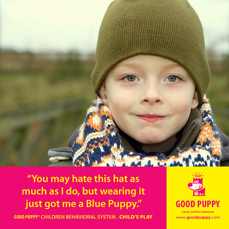 A young boy wearing a green beanie and a colorful scarf smiles subtly. The text overlay reads, "You may hate this hat as much as I do, but wearing it just got me a Blue Puppy." The image is promoting the Good Puppy Children Behavioral System, emphasizing the rewards system. The background is blurred, highlighting the boy's expression and outfit.