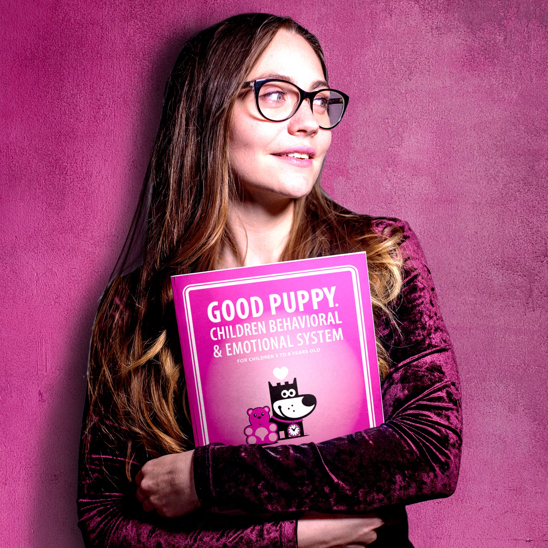 A woman with long hair and glasses is smiling while holding the "Good Puppy Children Behavioral & Emotional System" Therapy Manual & Toolkit against a pinkish-purple background. The manual is bright pink and designed for children aged 3 to 9. The woman is wearing a dark velvet-like top and appears content and confident, highlighting the appeal and professional use of the manual.